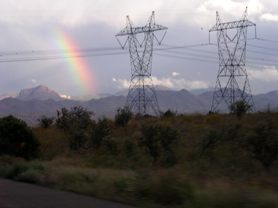 [In between the shrub grass in the foreground and the mountains in the distant are two high powerline towers. To the left of the towers is a vibrant splash of rainbow extending from the mountain tops to white clouds in the sky above on a leftward slant. From left to right one can easily see green, yellow and red. The violet at the far left blends with the dark sky color.]